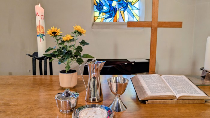Auf dem Tisch stehen eine Vase mit Sonnenblumen, silberne Kelche und ein offenes Buch neben einem Holzkreuz