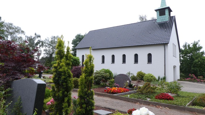 Weiße Kirche mit grünem Turm und Friedhof mit Grabsteinen und Blumenbeeten