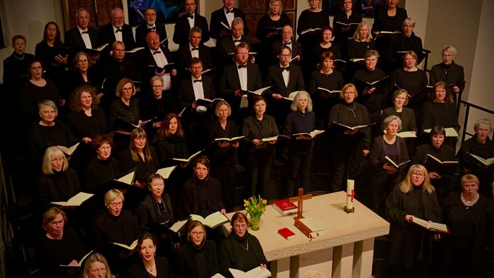 Ein großer Chor singt in einem Raum, alle in schwarzen Roben und mit Notenblättern.