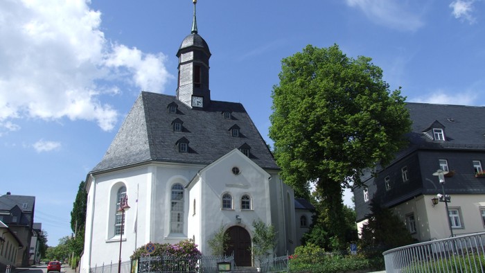 Weiße Kirche mit hohem Turm und Uhr, umgeben von Bäumen und Häusern