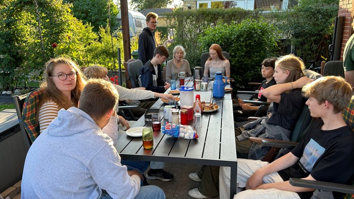 En gruppe af unge sidder udendørs ved en lang bord og spiser.
