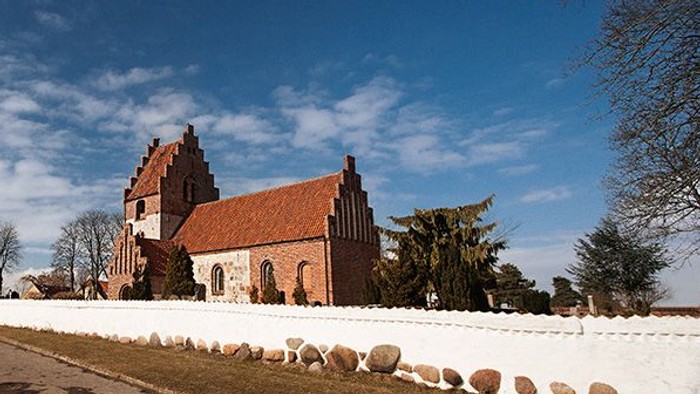 En rød kirke med spisse tårne og hvide murer, omgivet af træer og en klar blå himmel.