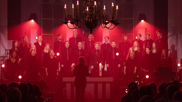 Ein Chor singt auf einer Bühne bei einem Konzert, beleuchtet von roten Scheinwerfern und einem großen Kronleuchter.