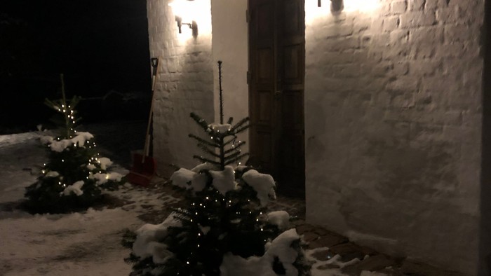 En snedækket juletree foran hvidkalket hus med lys.