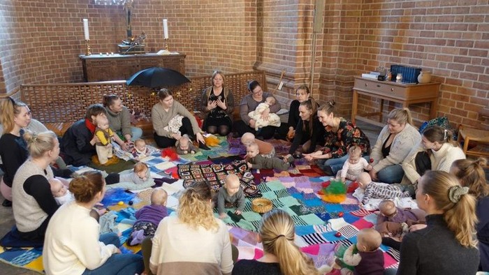 En gruppe af kvinder og børn sidder i en kreds og laver håndarbejde i et rum med mursten og lysende vinduer