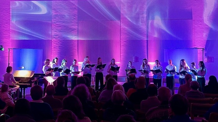 Et kor optræder på scenen under levende lilla belysning med et publikum siddende foran.