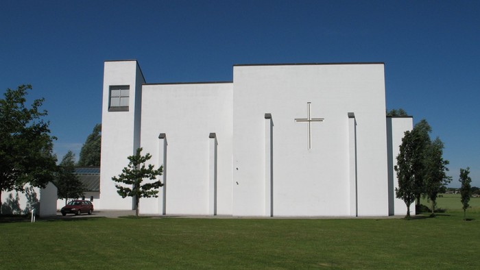 Moderne hvid kirke med korssymbol på facaden, omgivet af grøn græsplæne og træer.