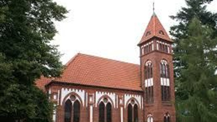 Das Bild zeigt eine rote Backsteinkirche mit hohem Turm und spitzen Fenstern, umgeben von Bäumen.