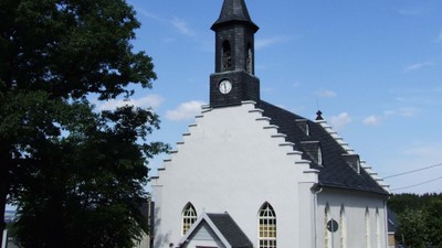 Kleine weiße Kirche mit schwarzem Turm und Uhr, umgeben von Bäumen und Büschen.