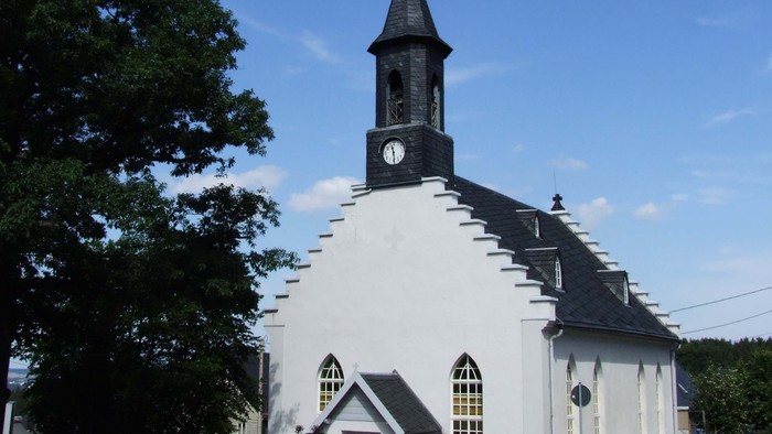 Kleine weiße Kirche mit schwarzem Turm und Uhr, umgeben von Bäumen und Büschen.