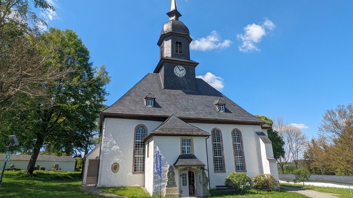 Weiße Kirche mit hohem Turm und Uhr, umgeben von Bäumen und Gras