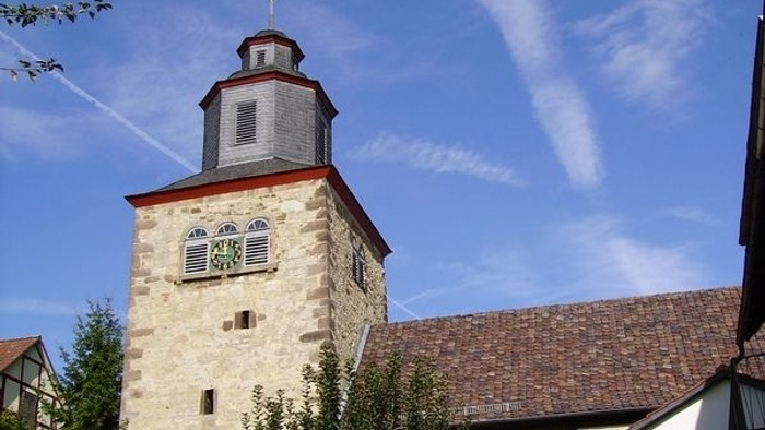 Steinerner Turm mit Uhr und Wetterfahne, umgeben von Gebäuden und Grünflächen unter blauem Himmel.