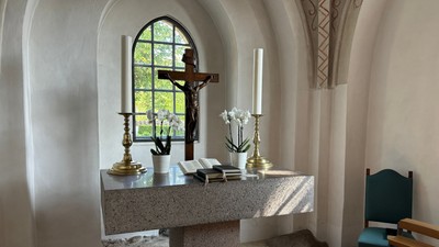 Roligt kapelalter med krucifiks, lys, blomster og bøger under et farvet glasmosaikvindue med bue.