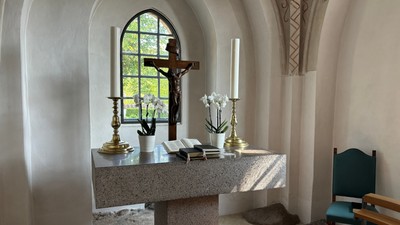 Roligt kapelalter med krucifiks, lys, blomster og bøger under et farvet glasmosaikvindue med bue.