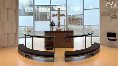 Moderne kapelinteriør med buede siddepladser, alter og store vinduer, der viser en rolig udsigt til naturen og græske bogstaver.