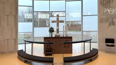 Moderne kapelinteriør med buede siddepladser, alter og store vinduer, der viser en rolig udsigt til naturen og græske bogstaver.