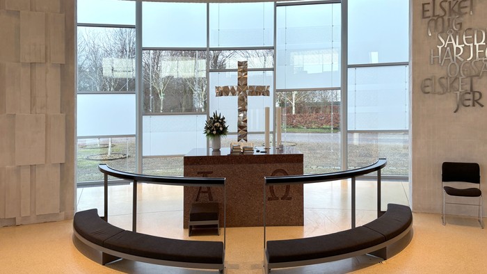 Moderne kapelinteriør med buede siddepladser, alter og store vinduer, der viser en rolig udsigt til naturen og græske bogstaver.