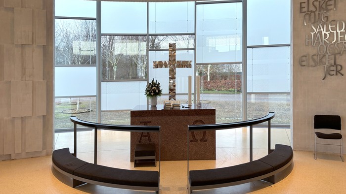 Moderne kapelinteriør med buede siddepladser, alter og store vinduer, der viser en rolig udsigt til naturen og græske bogstaver.