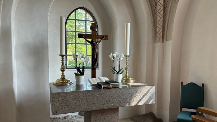 Roligt kapelalter med krucifiks, lys, blomster og bøger under et farvet glasmosaikvindue med bue.