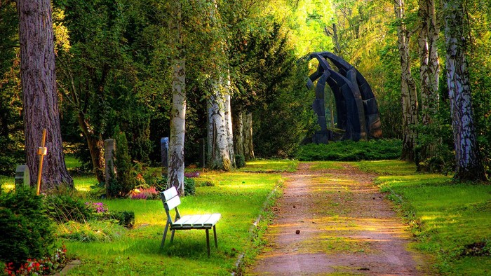 Ein Parkweg mit Bank und großer Holzskulptur umgeben von Bäumen