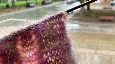 En person sitter ved vinduet og stråler en lilla trøje. Ude regner det og der er træer og en bil på vejen.