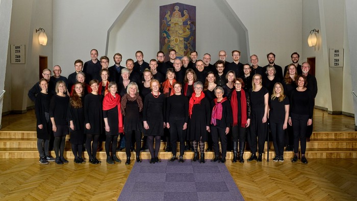 Et stort kor i ensartet sort tøj poserer sammen indendørs til et gruppefoto.