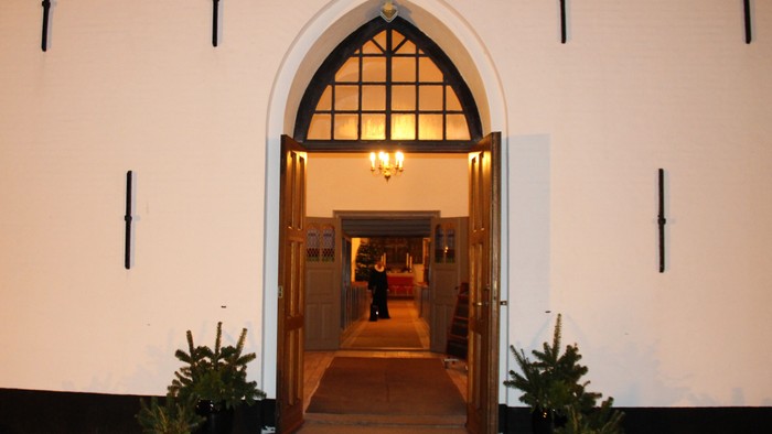 Indgangsdøren til Nr. Løgum kirke står åben, og ved hver side af døren står et par krukker pyntet med gran. Bagerst i kirken, på gangen, ses en præst i præstekjolen.