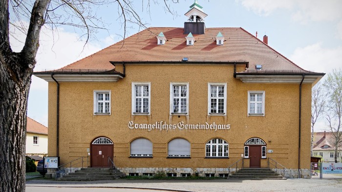 Frontalansicht vom Gemeindehaus Kreuz. Zweigeschossiger Bau mit ockerfarbener Fassade, Walmdach und kleinem mittigen Türmchen mit Glocke.