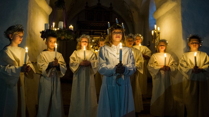 En gruppe kvinder i hvide kler og kranser holder lys i en kirke.