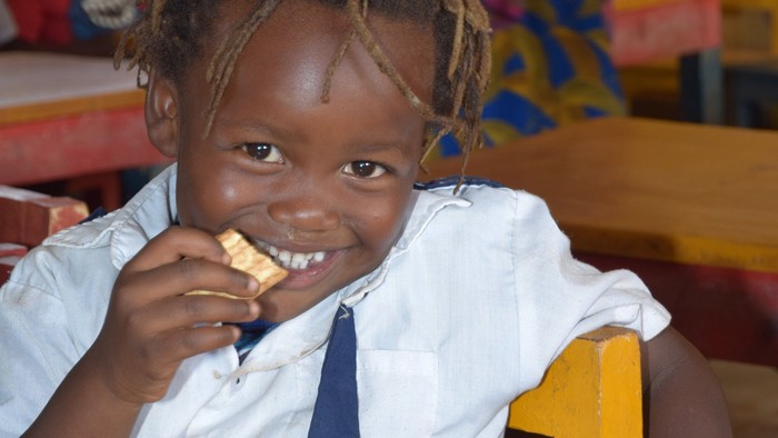 Et gladt lille barn i skoleuniform, der spiser en snack ved et gult skrivebord.