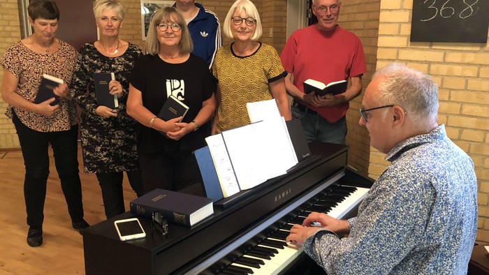 En gruppe af voksne står om et piano, hvor en mand spiller og de andre holder partiturer