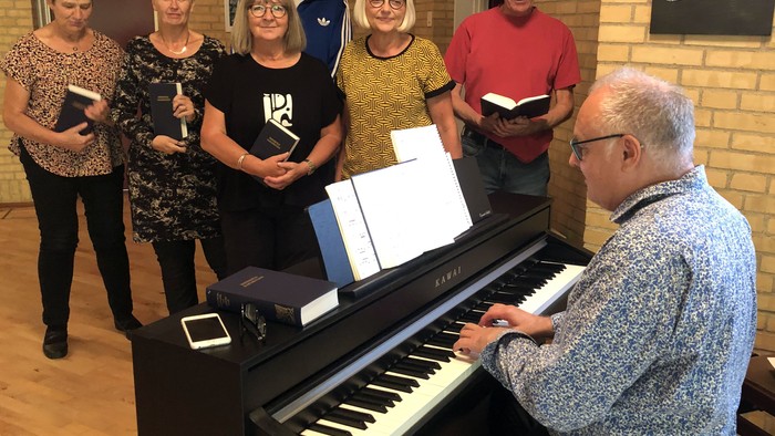 En gruppe af voksne står om et piano, hvor en mand spiller og de andre holder partiturer