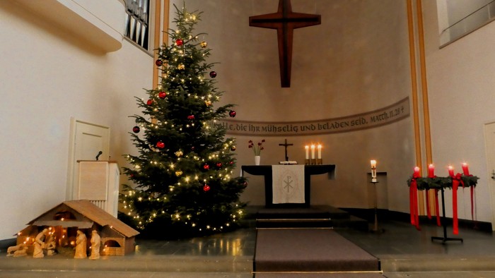 In der Kirche steht ein geschmückter Weihnachtsbaum neben einem Krippenspiel und einem Altar mit Kerzen.
