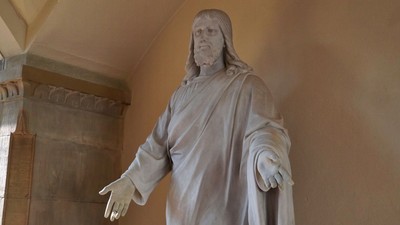 Eine Statue einer verhüllten Figur mit ausgebreiteten Armen vor einem strukturierten Wandhintergrund.