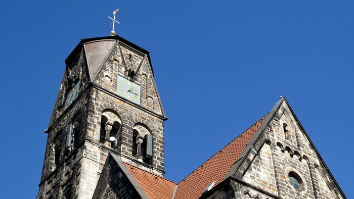 Historische Steinkirche mit hohem Glockenturm und steilem, rotziegelgedecktem Dach vor klarem blauem Himmel.