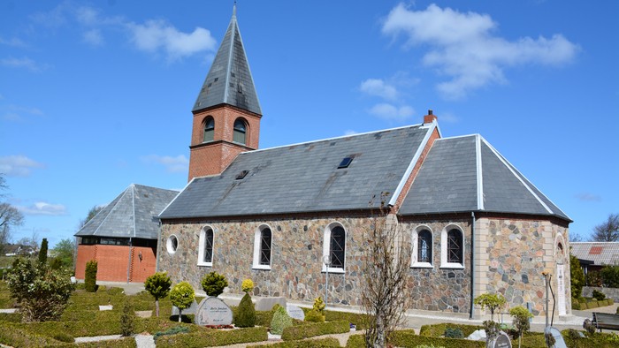 Hyggelig stenkirke med et højt spir omgivet af en velplejet kirkegård med klippede hække og grave.