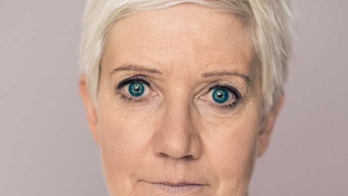 En person med kort, hvidt hår og iøjnefaldende blå øjne iført en lys bluse.