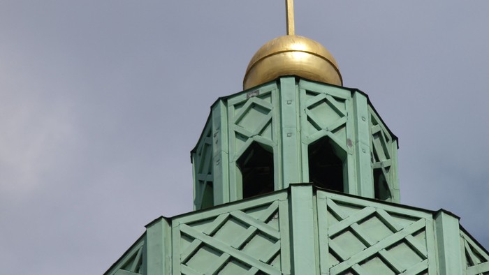 Das Bild zeigt einen grünen Glockenturm mit goldenem Kreuz und Kuppel.