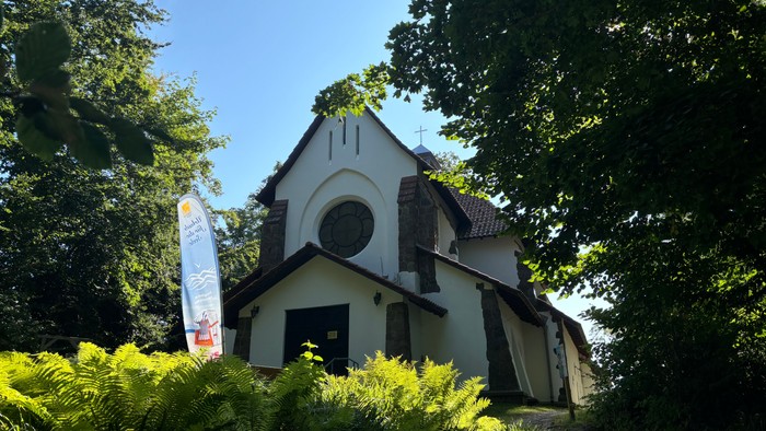 Kleine weiße Kirche mit Rundbogenfenster und Kreuz, umgeben von Bäumen und Büschen