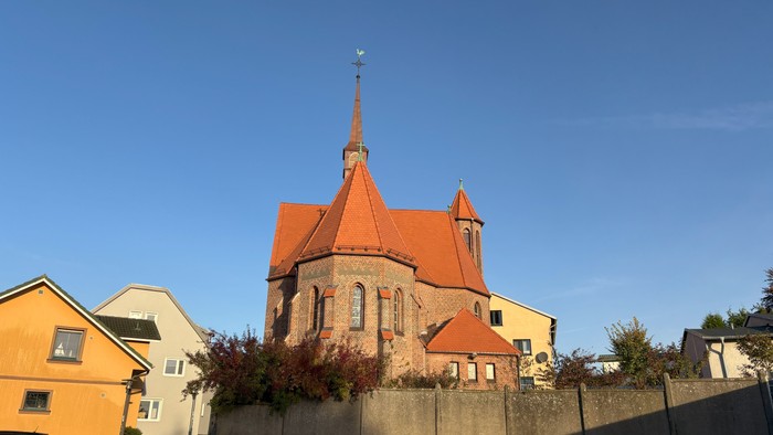 Kirchengebäude mit rotem Dach und spitzem Turm neben gelben Häusern