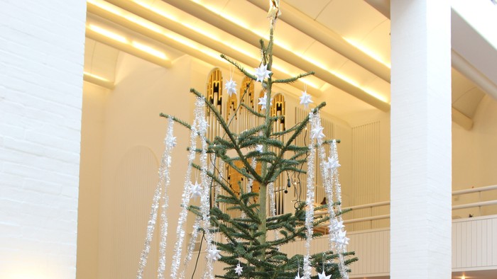 Et højt juletræ pyntet med hvide dekorationer og lys står i et rummeligt rum.