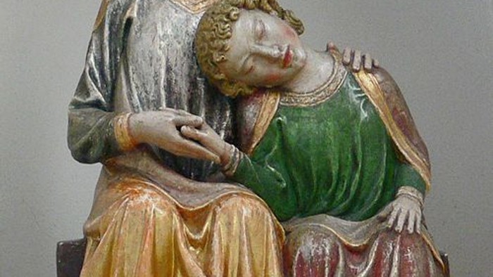 Holzstatue, die einen bärtigen Mann darstellt, der ein schlafendes Kind in traditionellen Gewändern zärtlich hält.