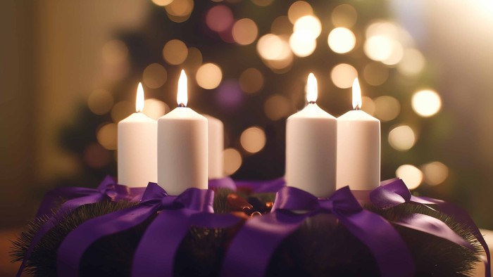 Fire tændte stearinlys i en dekorativ krans med lilla sløjfer, placeret mod en sløret baggrund med julelys.