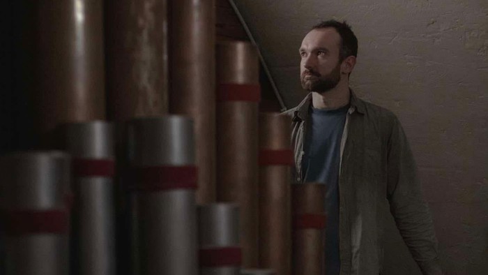 En mand står eftertænksomt mellem rækker af stablede tønder i et svagt oplyst rum.