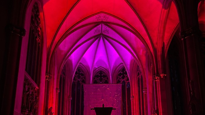 Bild des Chorraumes in St. Pankratius in der Fastenzeit. Das große Kreuz, das von der Decke hängt, ist verhüllt und der Chorraum ist in rotes und violettes Licht getaucht.