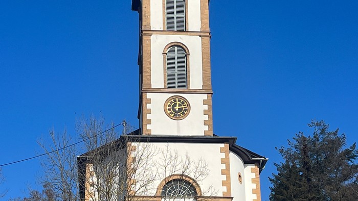 Kirchturm mit Uhr und Kreuz auf dem Dach, umgeben von Bäumen und Himmel