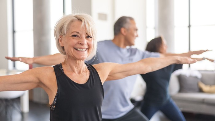 Eine ältere Frau strahlt während eines Fitnesskurses mit ausgebreiteten Armen.