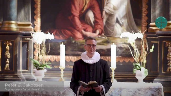 Mand i svart kapittel i kirke med bok og blomsterstykke.