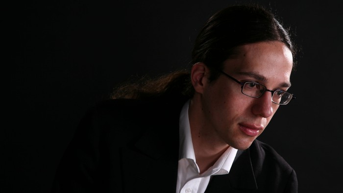 Ein nachdenklicher Mann mit Brille und langen Haaren in einem dunklen, stimmungsvollen Porträt.