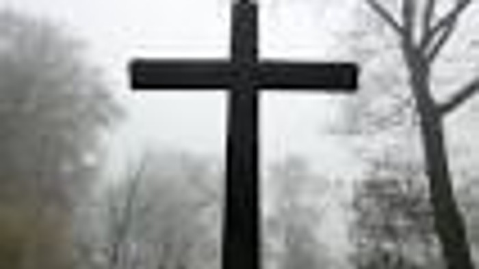 Zwei Kreuze vor bewölktem Himmel und Bäumen.